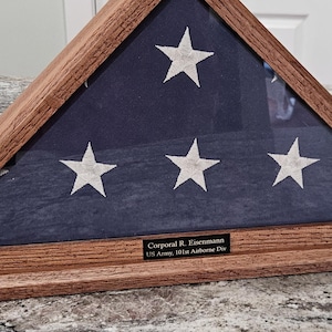 Puede incluir: Un estuche triangular de madera que contiene una bandera estadounidense plegada. La bandera es azul con estrellas y rayas blancas. Una pequeña placa con texto es visible en la parte inferior. La caja es de madera marrón claro.