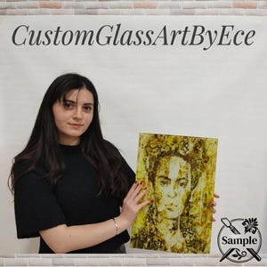 A woman in a black top is holding up a framed artwork of a woman's face, which appears to be a mosaic or collage.