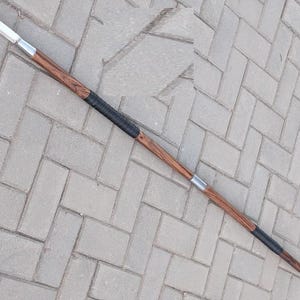 Puede incluir: Una lanza con un asta de madera y una punta de lanza de metal. El asta tiene un patrón de vetas de madera marrón oscuro con secciones envueltas en negro y bandas de metal plateado. La punta de lanza es de color plata claro.