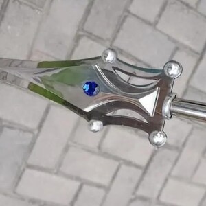 May include: A silver-colored sword with a decorative handle. The handle features a blue gem and clear, round accents. The blade has a textured design. The sword is held against a brick background.