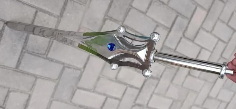 May include: A silver-colored sword with a decorative handle. The handle features a blue gem and clear, round accents. The blade has a textured design. The sword is held against a brick background.