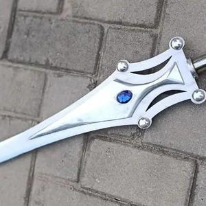 May include: A silver-colored sword with a decorative hilt featuring a blue gem. The blade is long and pointed, and the hilt has a stylized design with rounded accents. The sword is displayed on a brick surface.
