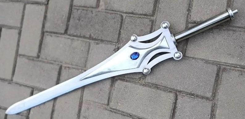 May include: A silver-colored sword with a decorative hilt featuring a blue gem. The blade is long and pointed, and the hilt has a stylized design with rounded accents. The sword is displayed on a brick surface.