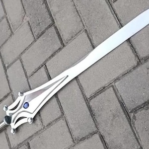 May include: A silver-colored sword with a long, pointed blade. The hilt features a decorative design with a blue gem in the center. The sword is lying on a gray brick surface.