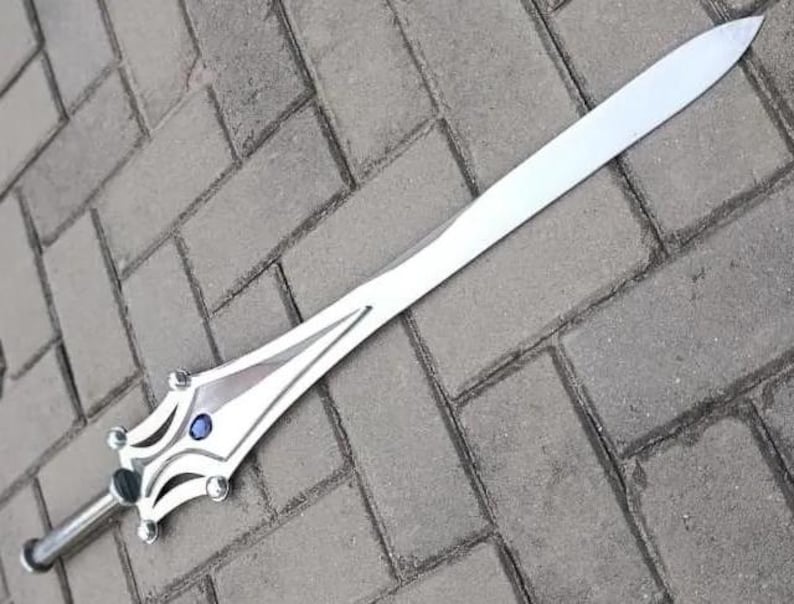 May include: A silver-colored sword with a long, pointed blade. The hilt features a decorative design with a blue gem in the center. The sword is lying on a gray brick surface.