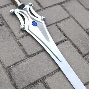 May include: A silver sword with a decorative hilt featuring a blue gem. The blade is long and straight, with a polished finish. The sword is lying on a gray brick surface.