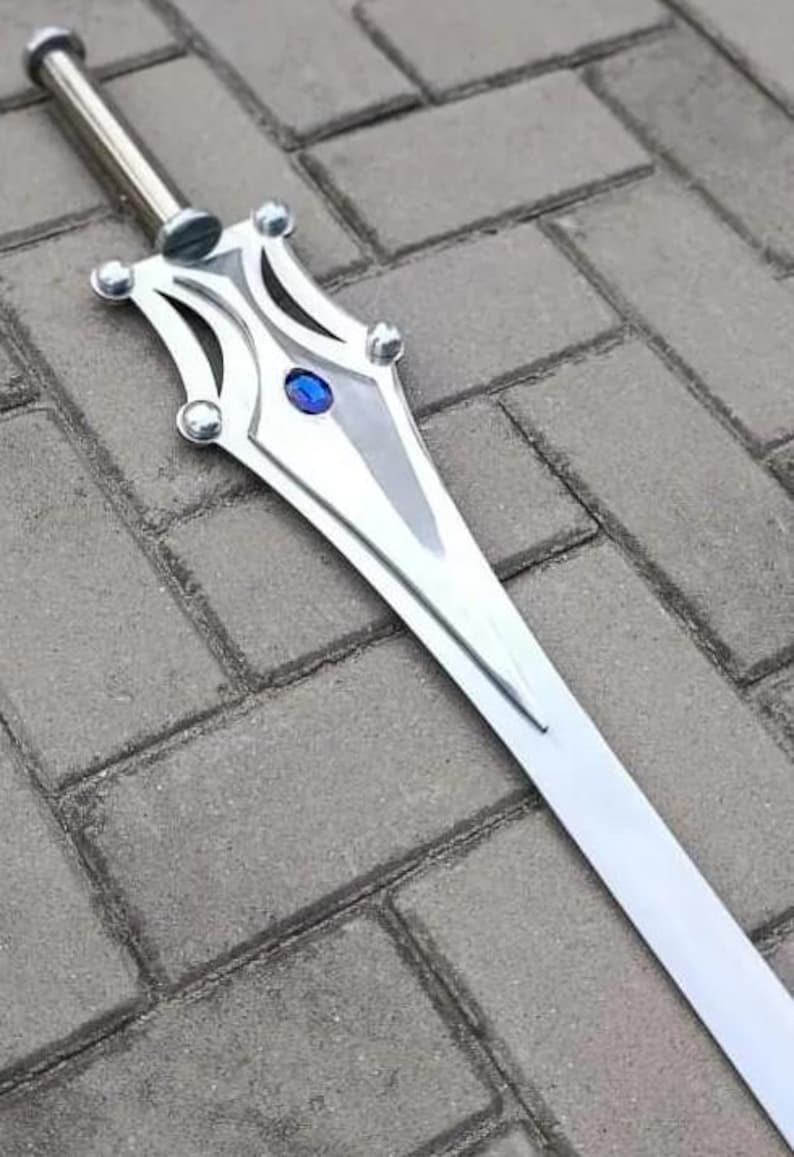 May include: A silver sword with a decorative hilt featuring a blue gem. The blade is long and straight, with a polished finish. The sword is lying on a gray brick surface.