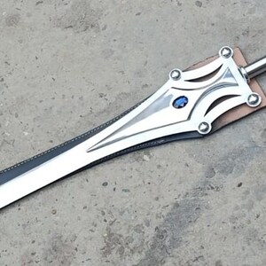 May include: A silver sword with a polished blade and ornate hilt. The hilt features a blue gem and decorative rivets. The sword rests on a brown leather base, with a black leather sheath. The overall length is approximately 30 inches.