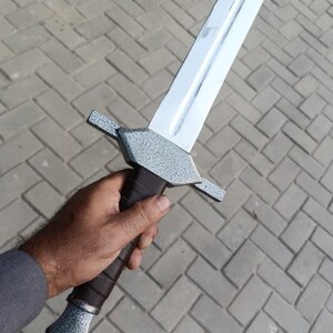 May include: A silver-bladed sword with a textured gray hilt and guard, held in a hand. The sword's blade is polished, reflecting light. The hilt is wrapped in dark brown leather. The guard has a textured, speckled finish.