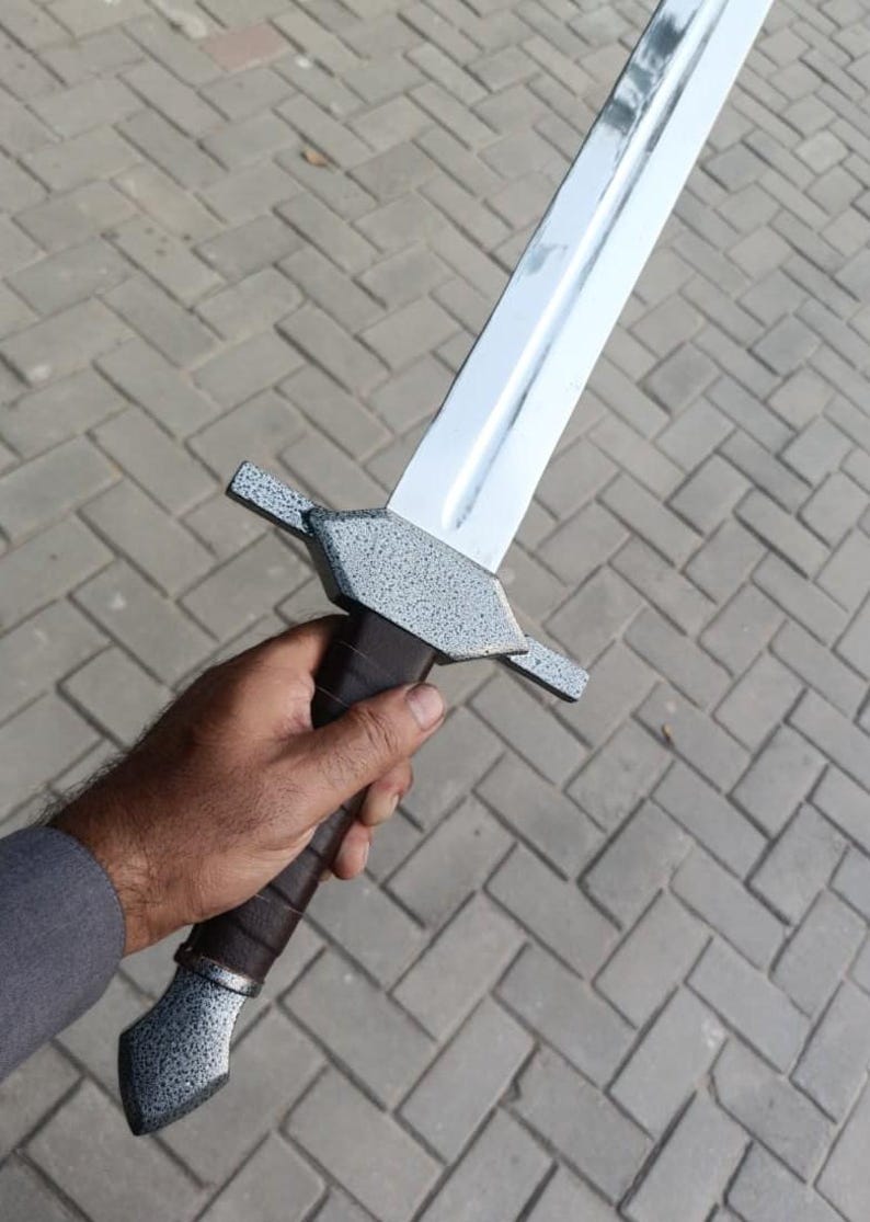 May include: A silver-bladed sword with a textured gray hilt and guard, held in a hand. The sword's blade is polished, reflecting light. The hilt is wrapped in dark brown leather. The guard has a textured, speckled finish.