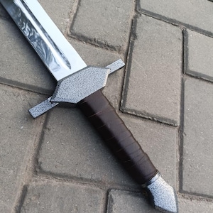 May include: A medieval-style sword with a polished silver blade and a textured gray crossguard. The handle is wrapped in dark brown leather. The sword is lying on a gray brick surface.