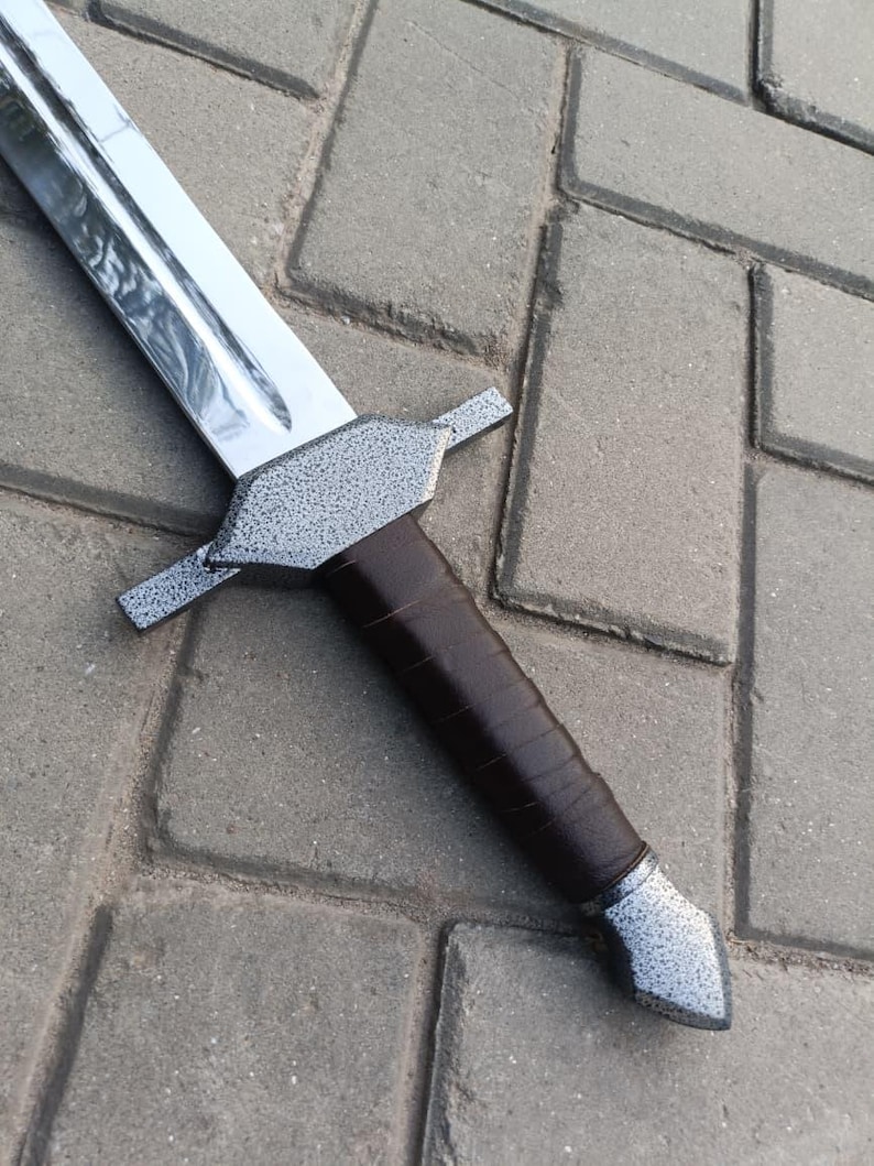 May include: A medieval-style sword with a polished silver blade and a textured gray crossguard. The handle is wrapped in dark brown leather. The sword is lying on a gray brick surface.