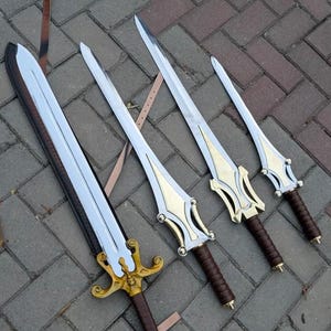 May include: Four swords with silver blades and gold and brown handles are displayed on a brick surface. The swords vary in size, with ornate gold detailing on the hilts and brown leather-wrapped grips. A leather strap is visible.