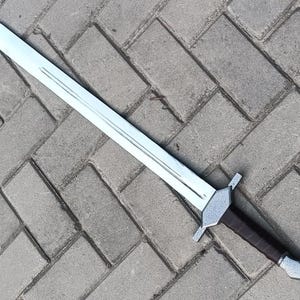 May include: A silver-colored sword with a long, straight blade and a brown handle. The sword has a crossguard and a pointed tip. The handle appears to be wrapped in leather. The sword is lying on a gray brick surface.