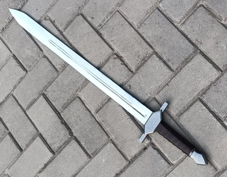 May include: A silver-colored sword with a long, straight blade and a brown handle. The sword has a crossguard and a pointed tip. The handle appears to be wrapped in leather. The sword is lying on a gray brick surface.