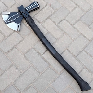 May include: A black and silver axe with a long, black wooden handle. The axe head features a silver blade with black accents. The handle is wrapped in black tape. The axe is lying on a gray brick surface.