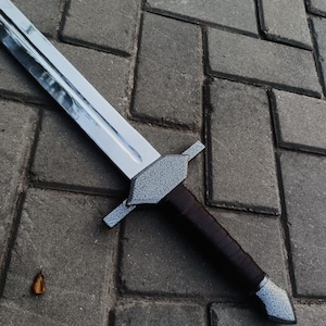 May include: A silver-bladed sword with a textured gray hilt and a dark brown leather-wrapped handle. The sword is lying on a gray brick surface.
