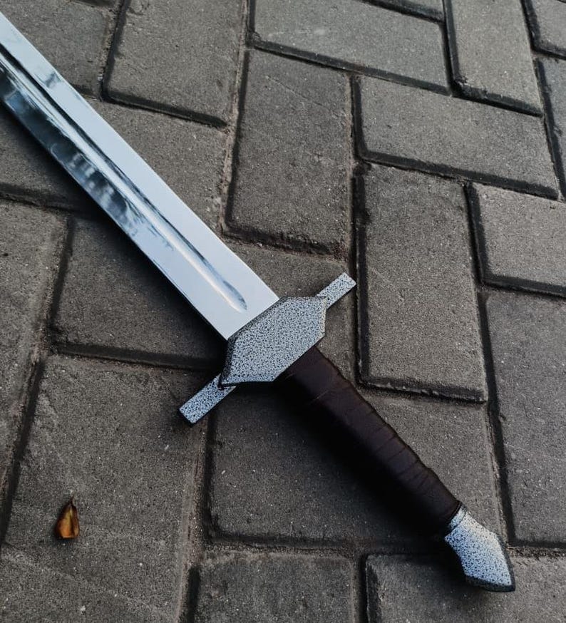 May include: A silver-bladed sword with a textured gray hilt and a dark brown leather-wrapped handle. The sword is lying on a gray brick surface.