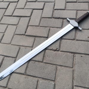 May include: A long, silver sword with a double-edged blade. The hilt features a brown leather-wrapped grip and a silver crossguard. The sword is lying on a gray brick surface.