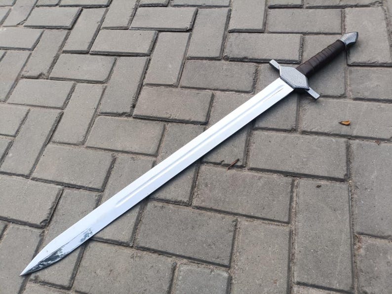 May include: A long, silver sword with a double-edged blade. The hilt features a brown leather-wrapped grip and a silver crossguard. The sword is lying on a gray brick surface.