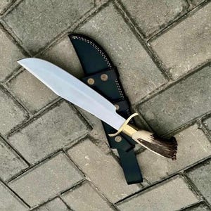 Peut inclure: Un grand couteau à lame argentée avec un manche en laiton et une poignée en corne marron, posé sur un étui en cuir noir. Le couteau a une large lame et un bord incurvé. L'étui a des rivets en laiton et des coutures blanches.