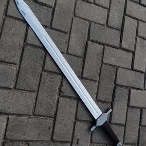 May include: A silver-colored sword with a long, straight blade and a dark brown handle. The sword has a crossguard and a pointed tip. The blade appears to be made of metal. The sword is lying on a gray brick surface.
