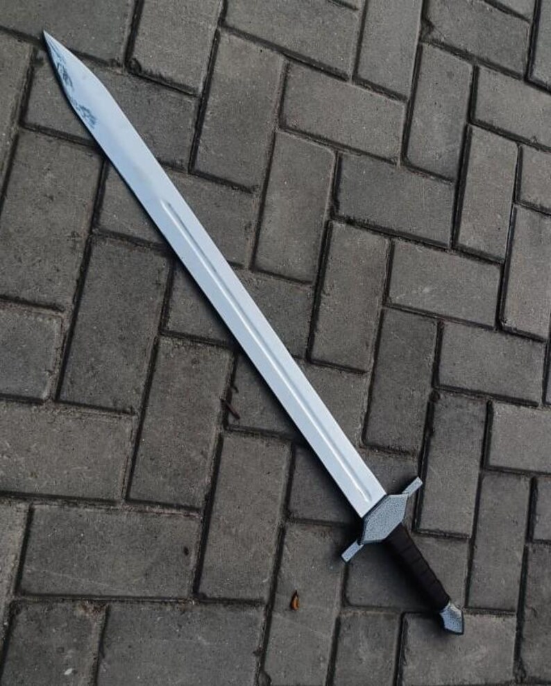 May include: A silver-colored sword with a long, straight blade and a dark brown handle. The sword has a crossguard and a pointed tip. The blade appears to be made of metal. The sword is lying on a gray brick surface.
