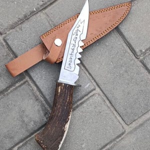Könnte beinhalten: Ein Jagdmesser mit gezahnter Klinge und einem Hirschhorngriff. Die Klinge hat geätzten Text und wird mit einer braunen Lederscheide geliefert. Das Messer ist für den Außeneinsatz konzipiert.