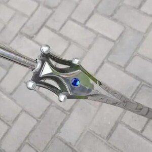 May include: A silver-colored sword with a decorative hilt and a blue gem in the center. The sword has a long, pointed blade and is lying on a gray brick surface. The hilt has a unique design with rounded accents.
