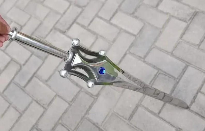 May include: A silver-colored sword with a decorative hilt and a blue gem in the center. The sword has a long, pointed blade and is lying on a gray brick surface. The hilt has a unique design with rounded accents.