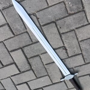 May include: A long, silver-colored sword with a pointed tip and a brown handle. The sword has a straight blade and a silver crossguard. The handle is wrapped in a dark brown material. The sword is lying on a gray brick surface.
