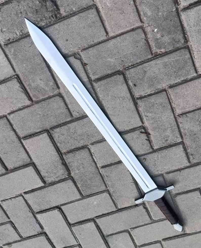 May include: A long, silver-colored sword with a pointed tip and a brown handle. The sword has a straight blade and a silver crossguard. The handle is wrapped in a dark brown material. The sword is lying on a gray brick surface.