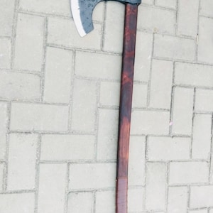 May include: A large, hand-forged axe with a dark, weathered steel blade and a long, stained wooden handle. The handle is wrapped with a leather grip near the base. The axe is designed for heavy-duty use.