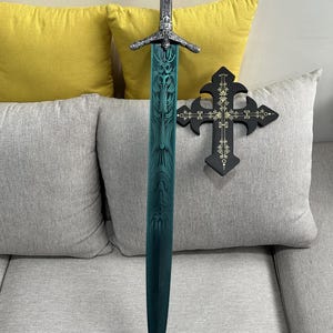May include: A decorative sword with a teal blade and ornate silver hilt. A black cross with gold accents is to the right. The sword is approximately 102 cm long. The items are displayed on a gray couch with yellow cushions.