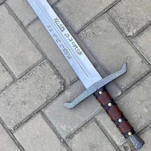 以下が含まれることがあります： 両刃の長い銀色の剣。柄は金色のスタッドと黒のアクセントが付いた茶色の革で包まれています。ガードとポメルはテクスチャ加工されたグレー仕上げです。ブレードにはエッチングされたテキストがあります。