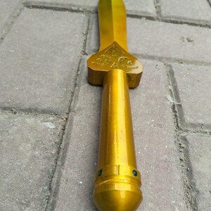 May include: A gold-coloured dagger with a pointed blade and a detailed hilt. The handle is cylindrical with two small green gems near the base. The dagger is made of a shiny, metallic material.