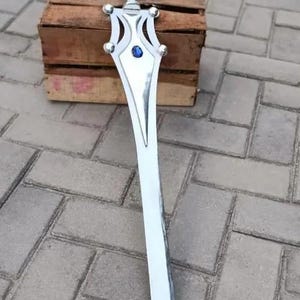 May include: A silver-colored sword with a decorative hilt and a blue gem in the center. The sword is resting on a wooden crate and is set against a gray brick background. The blade is long and pointed.