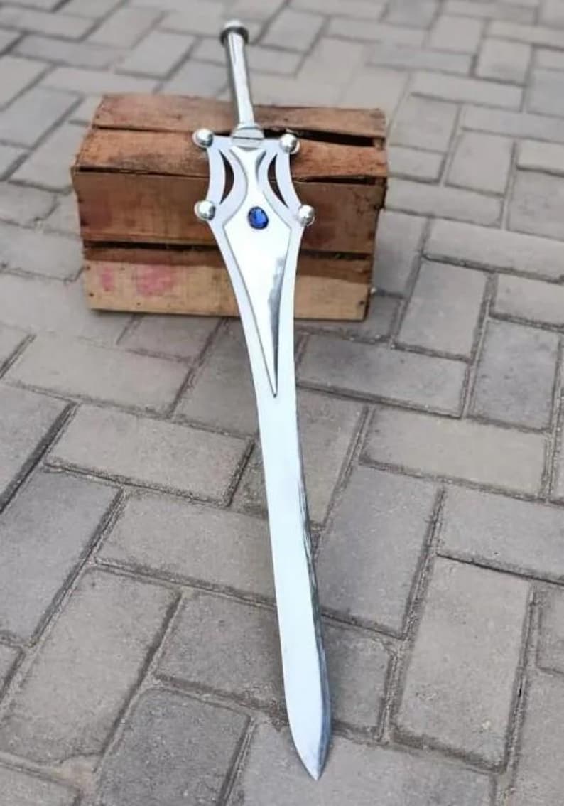 May include: A silver-colored sword with a decorative hilt and a blue gem in the center. The sword is resting on a wooden crate and is set against a gray brick background. The blade is long and pointed.