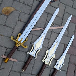 May include: Four fantasy swords with silver blades and ornate gold and brown handles. The largest sword has a brown leather sheath. The swords are arranged diagonally on a gray brick surface.