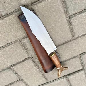 May include: A large, handmade knife with a polished steel blade and a wooden handle. The handle features a brass bolster and a decorative antler-shaped end. The knife rests in a brown leather sheath, set against a gray brick background.