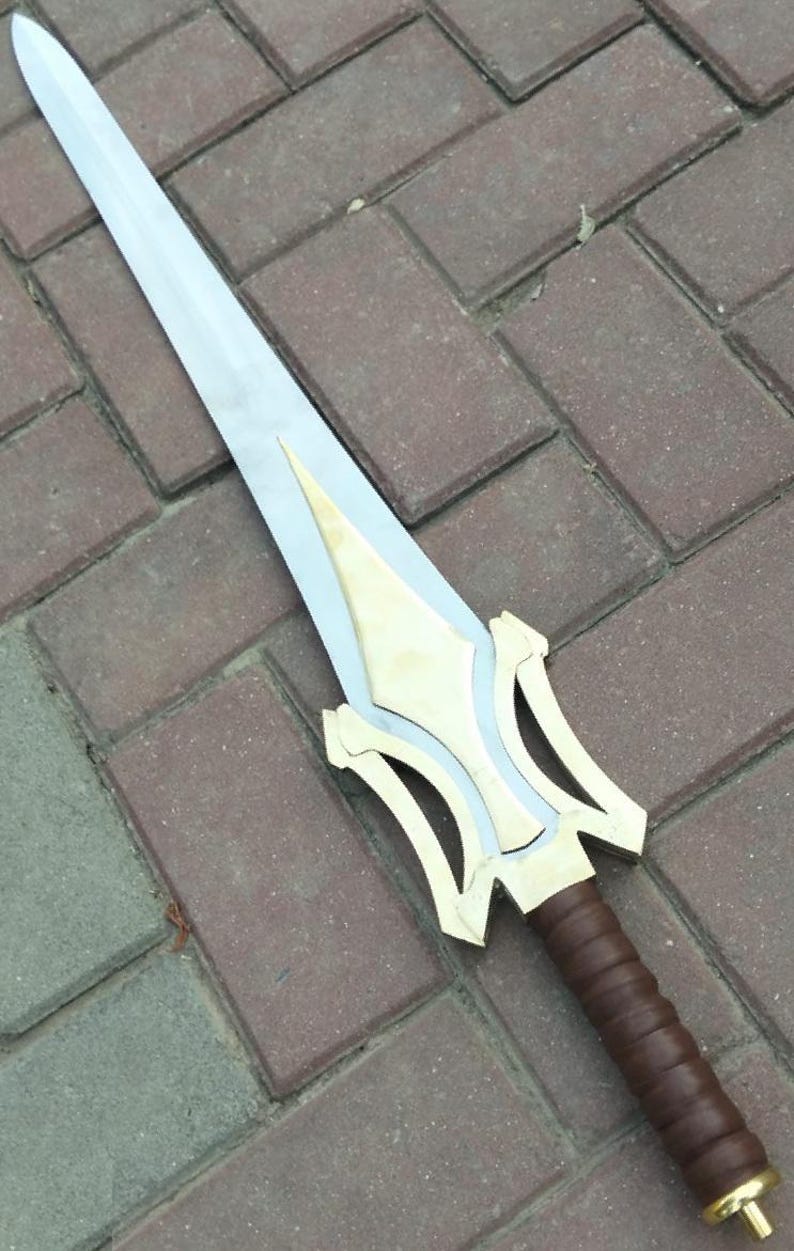Może przedstawiać: Srebrny miecz ze złotą rękojeścią i akcentami. Rękojeść jest owinięta brązową sk&oacute;rą. Miecz leży na ceglanej powierzchni. Miecz jest rekwizytem lub przedmiotem dekoracyjnym.