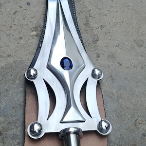 May include: A polished metal spearhead with a central blue gem and decorative cutouts. The spearhead is attached to a leather backing and a cylindrical metal shaft. The design is symmetrical and detailed.