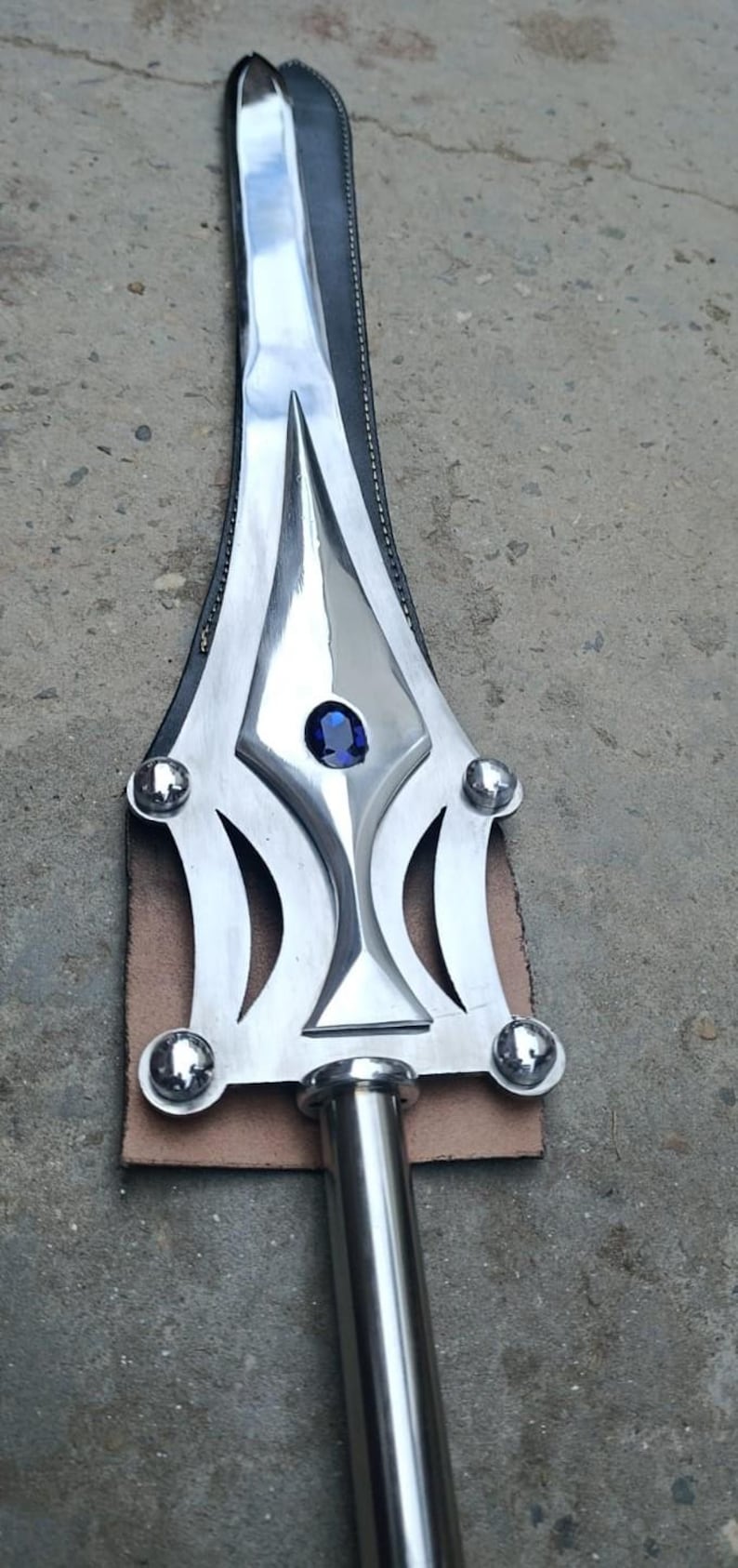 May include: A polished metal spearhead with a central blue gem and decorative cutouts. The spearhead is attached to a leather backing and a cylindrical metal shaft. The design is symmetrical and detailed.