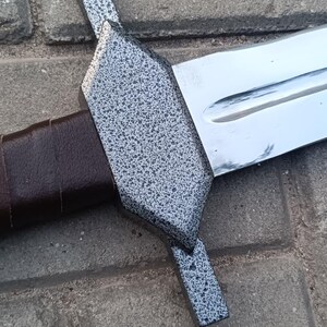 May include: A close-up of a sword with a silver blade and a textured, speckled gray crossguard. The handle is wrapped in brown leather. The sword is resting on a brick surface.