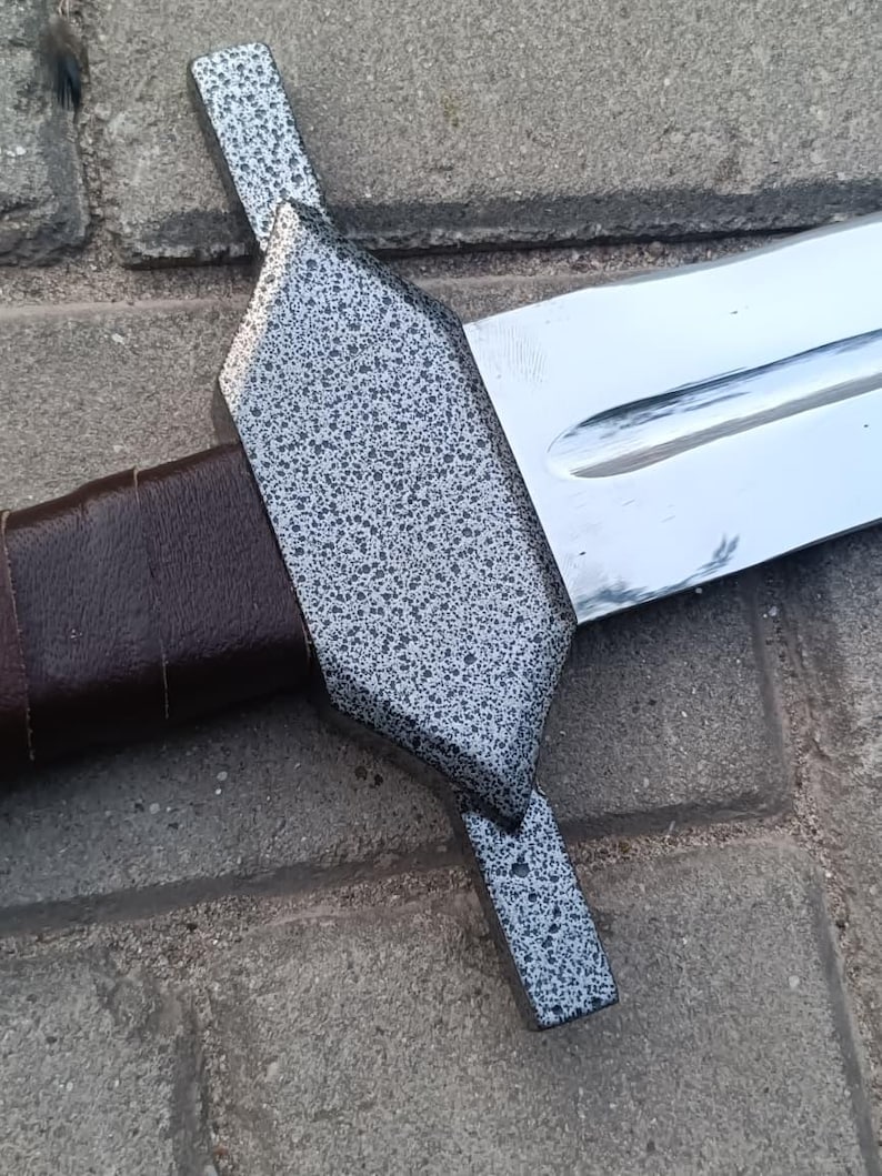 May include: A close-up of a sword with a silver blade and a textured, speckled gray crossguard. The handle is wrapped in brown leather. The sword is resting on a brick surface.