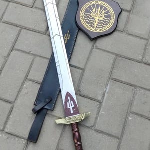 May include: A decorative sword with a white blade and a brown and gold handle, resting on a black sheath. A matching shield with a gold trident design is also present. The sword and shield are likely part of a costume or display set.