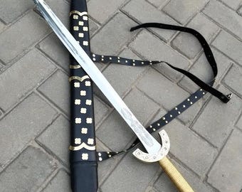 Puede incluir: Una espada decorativa con una hoja plateada y un mango dorado. La espada se muestra con su vaina negra, que tiene detalles dorados. También se incluye una correa de cuero negro.