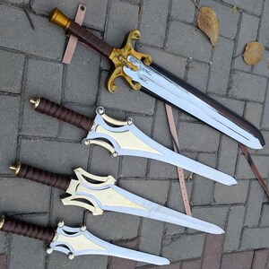 May include: An overhead shot of four fantasy swords. The swords have silver blades, gold and silver crossguards, and brown leather-wrapped handles. One sword is in a sheath with a brown leather strap. The swords are lying on a gray brick surface.