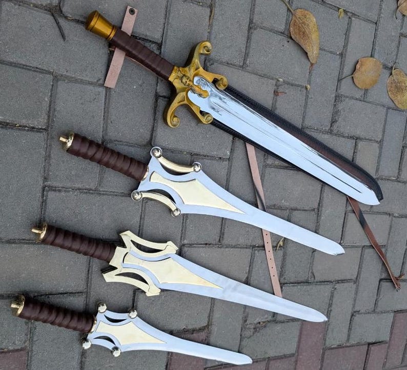 May include: An overhead shot of four fantasy swords. The swords have silver blades, gold and silver crossguards, and brown leather-wrapped handles. One sword is in a sheath with a brown leather strap. The swords are lying on a gray brick surface.