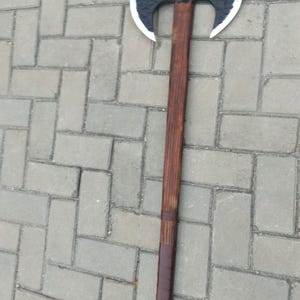 May include: A double-bladed axe with a dark, textured head and a polished silver edge. The long wooden handle is stained a rich brown and wrapped with a darker leather grip. The axe is lying on a gray brick surface.